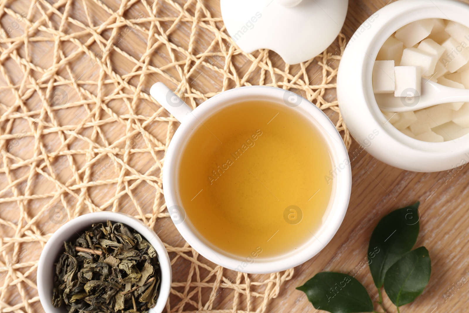 Photo of Refreshing green tea in cup, sugar bowl and leaves on wooden table, flat lay