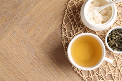 Photo of Refreshing green tea in cup, sugar bowl and dry leaves on wooden table, top view. Space for text