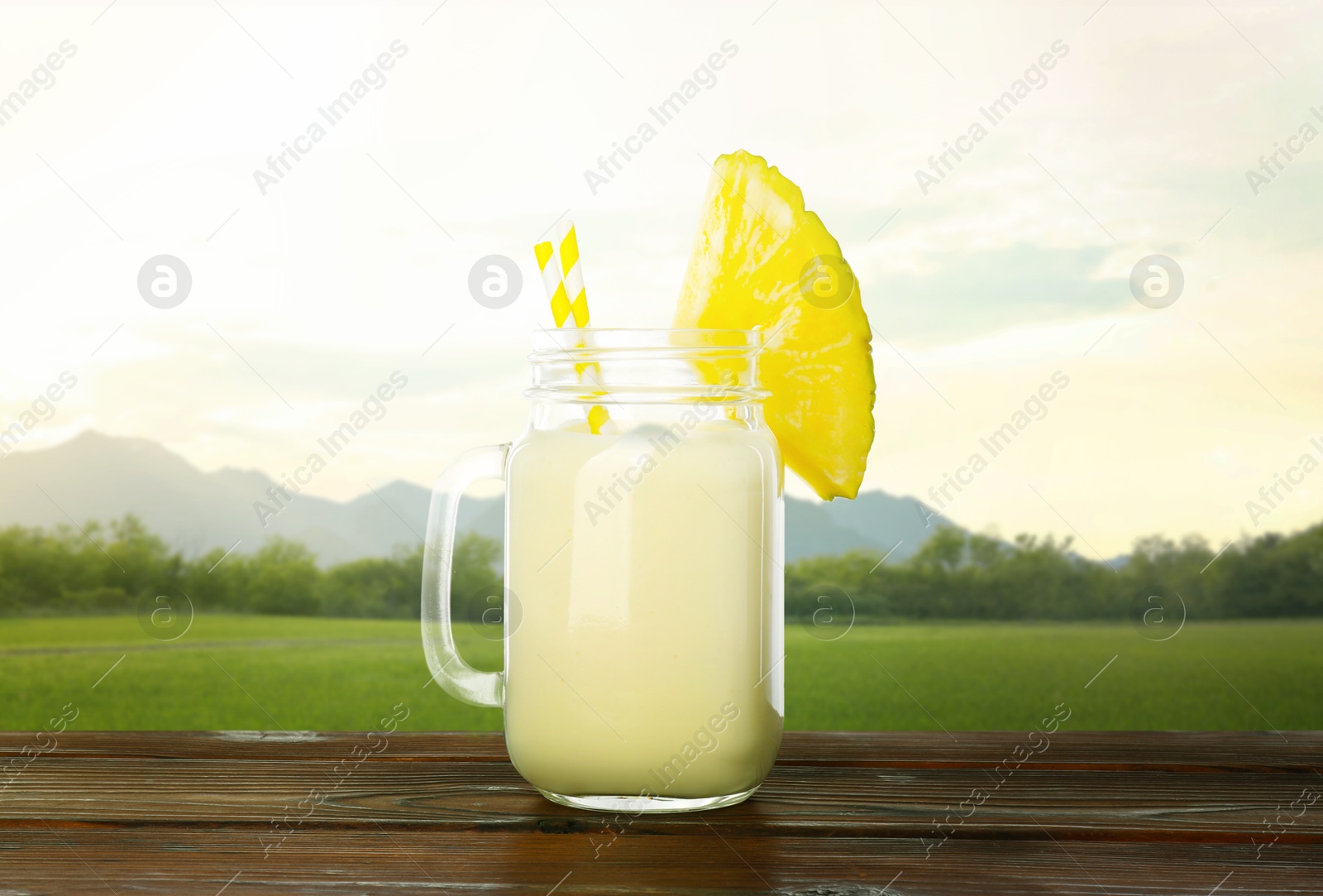 Image of Tasty pineapple cocktail or smoothie in mason jar on wooden table against mountain landscape