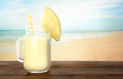 Image of Tasty pineapple cocktail or smoothie in mason jar on wooden table against seascape. Space for text