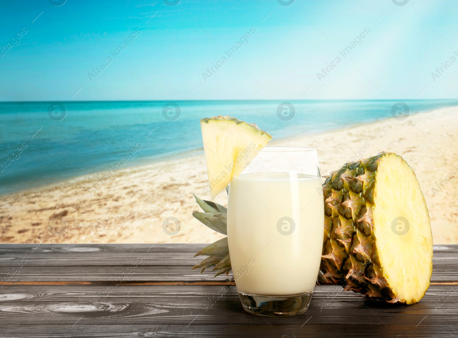 Image of Tasty pineapple cocktail or smoothie in glass and fresh fruit on wooden table against seascape. Space for text