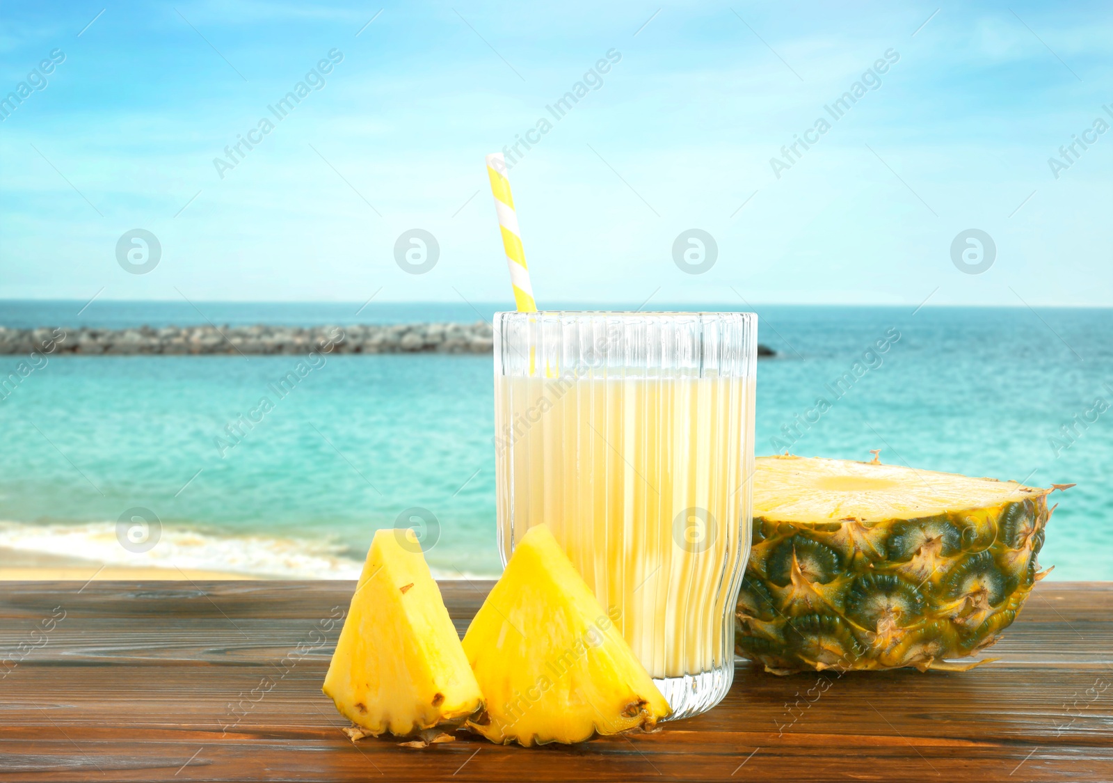 Image of Tasty pineapple cocktail or smoothie in glass and fresh fruit on wooden table against seascape