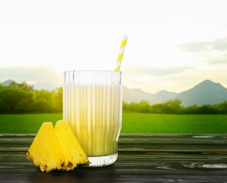 Image of Tasty pineapple cocktail or smoothie in glass and fresh fruit on wooden table against mountain landscape. Space for text