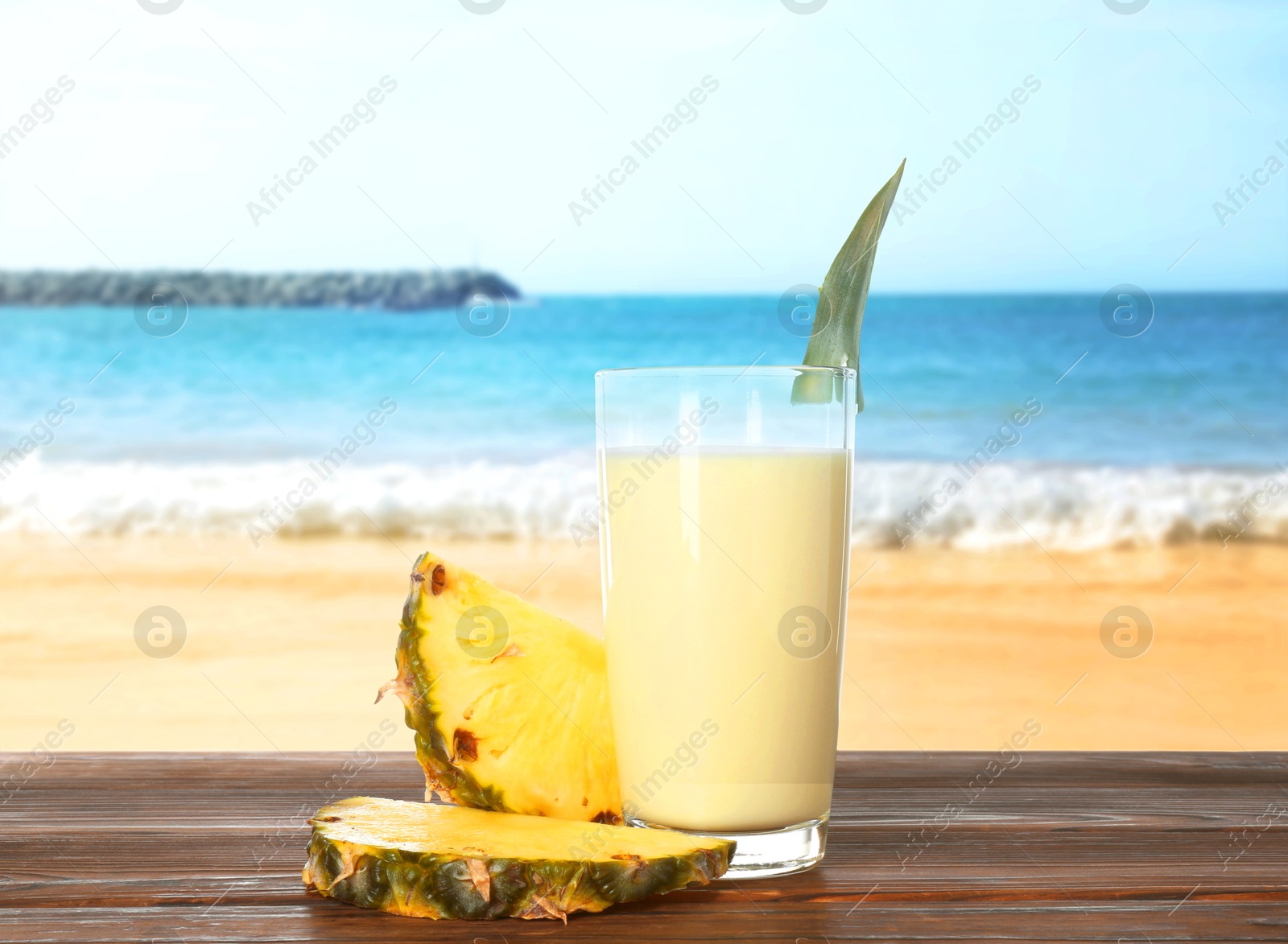 Image of Tasty pineapple cocktail or smoothie in glass and fresh fruit on wooden table against seascape