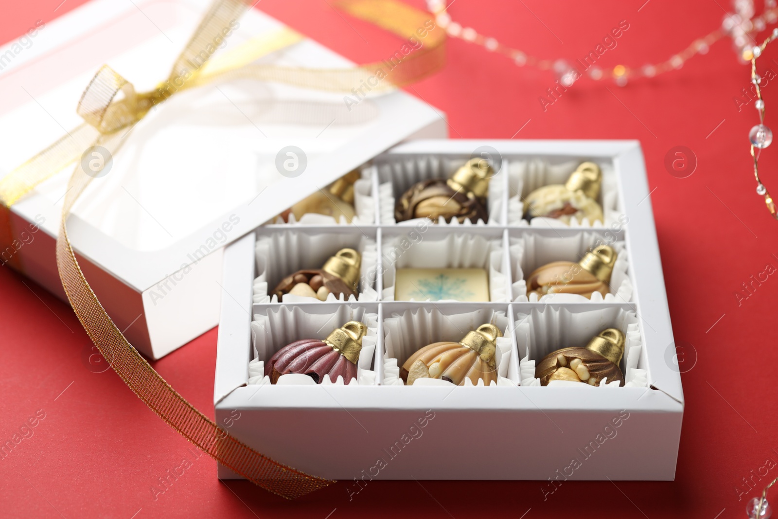 Photo of Box with tasty chocolate candies in shape of baubles and Christmas decor on red background, closeup