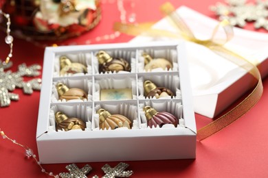 Photo of Box with tasty chocolate candies in shape of baubles and Christmas decor on red background, closeup