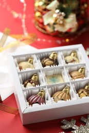 Photo of Box with tasty chocolate candies in shape of baubles and Christmas decor on red background, closeup