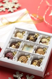 Photo of Box with tasty chocolate candies in shape of baubles and Christmas decor on red background, closeup