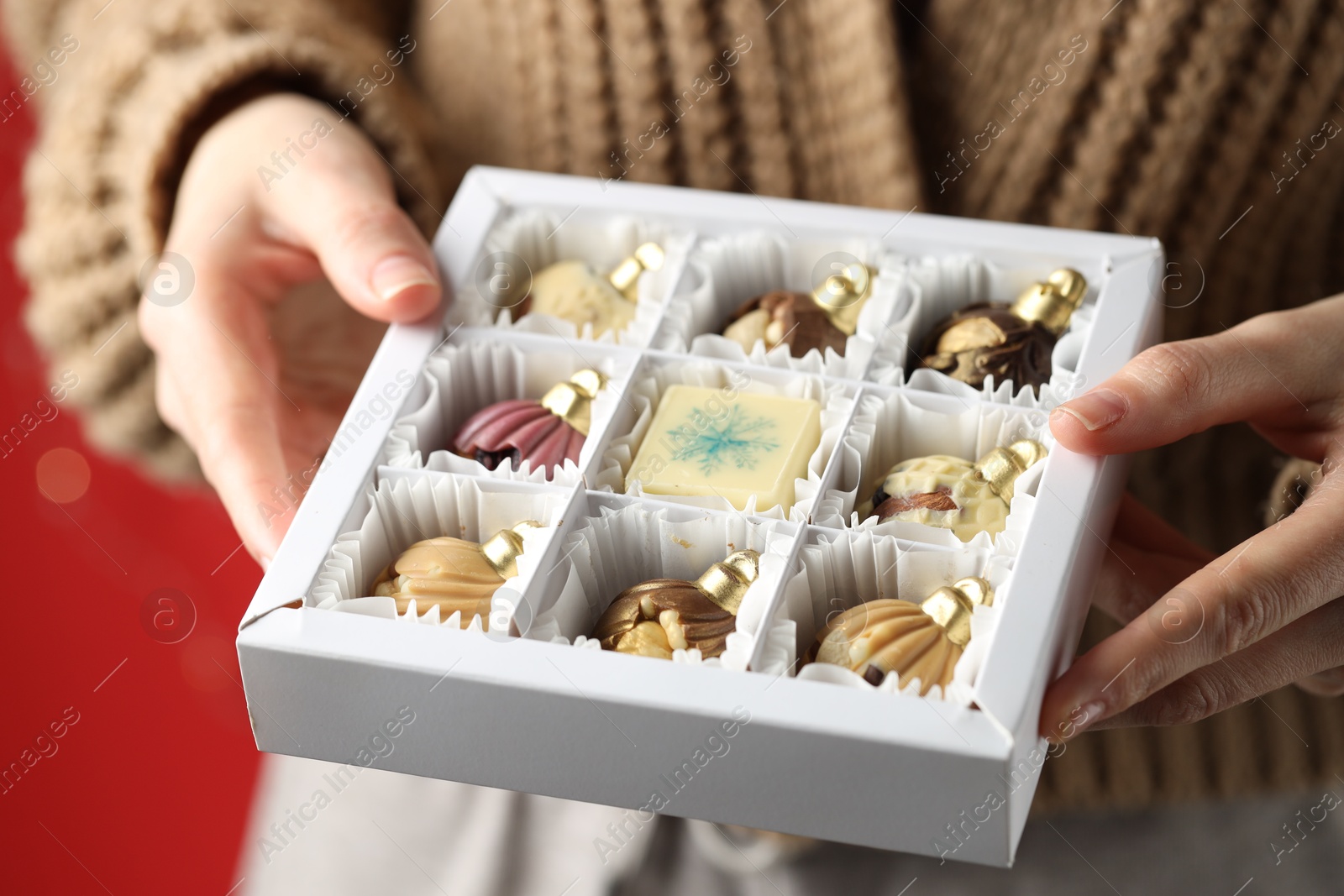 Photo of Woman holding box with tasty chocolate candies in shape of baubles on red background, closeup