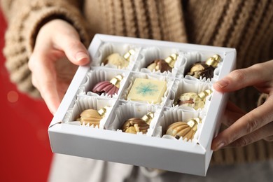 Photo of Woman holding box with tasty chocolate candies in shape of baubles on red background, closeup