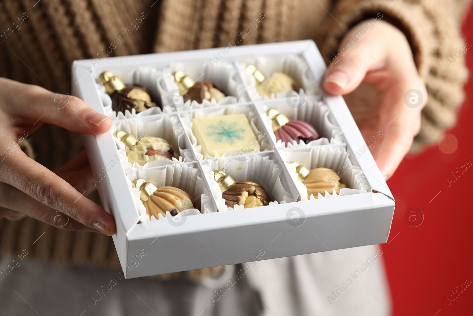 Photo of Woman holding box with tasty chocolate candies in shape of baubles on red background, closeup