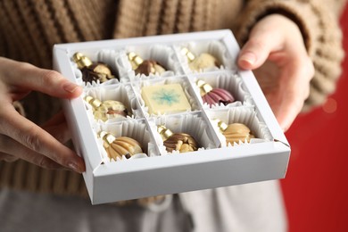 Photo of Woman holding box with tasty chocolate candies in shape of baubles on red background, closeup