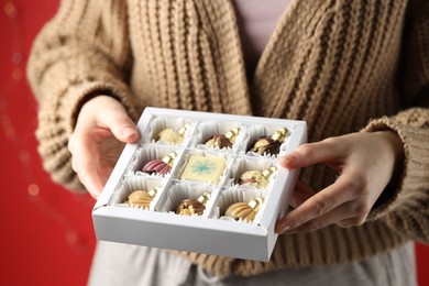 Photo of Woman holding box with tasty chocolate candies in shape of baubles on red background, closeup