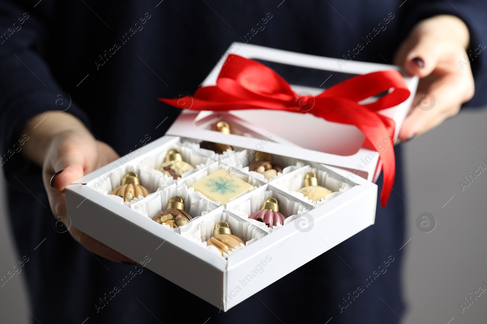 Photo of Woman holding box with tasty chocolate candies in shape of baubles on grey background, closeup