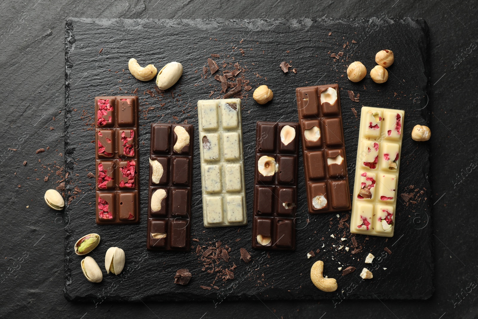 Photo of Different tasty chocolate bars with nuts and freeze dried raspberry on black table, flat lay