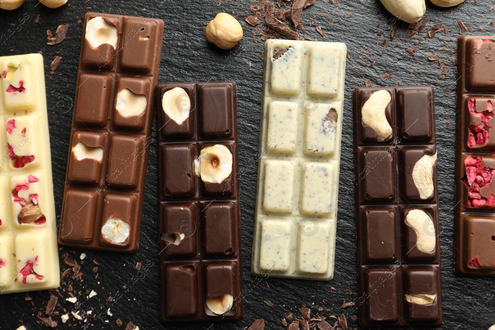 Photo of Different tasty chocolate bars with nuts and freeze dried raspberry on black table, flat lay