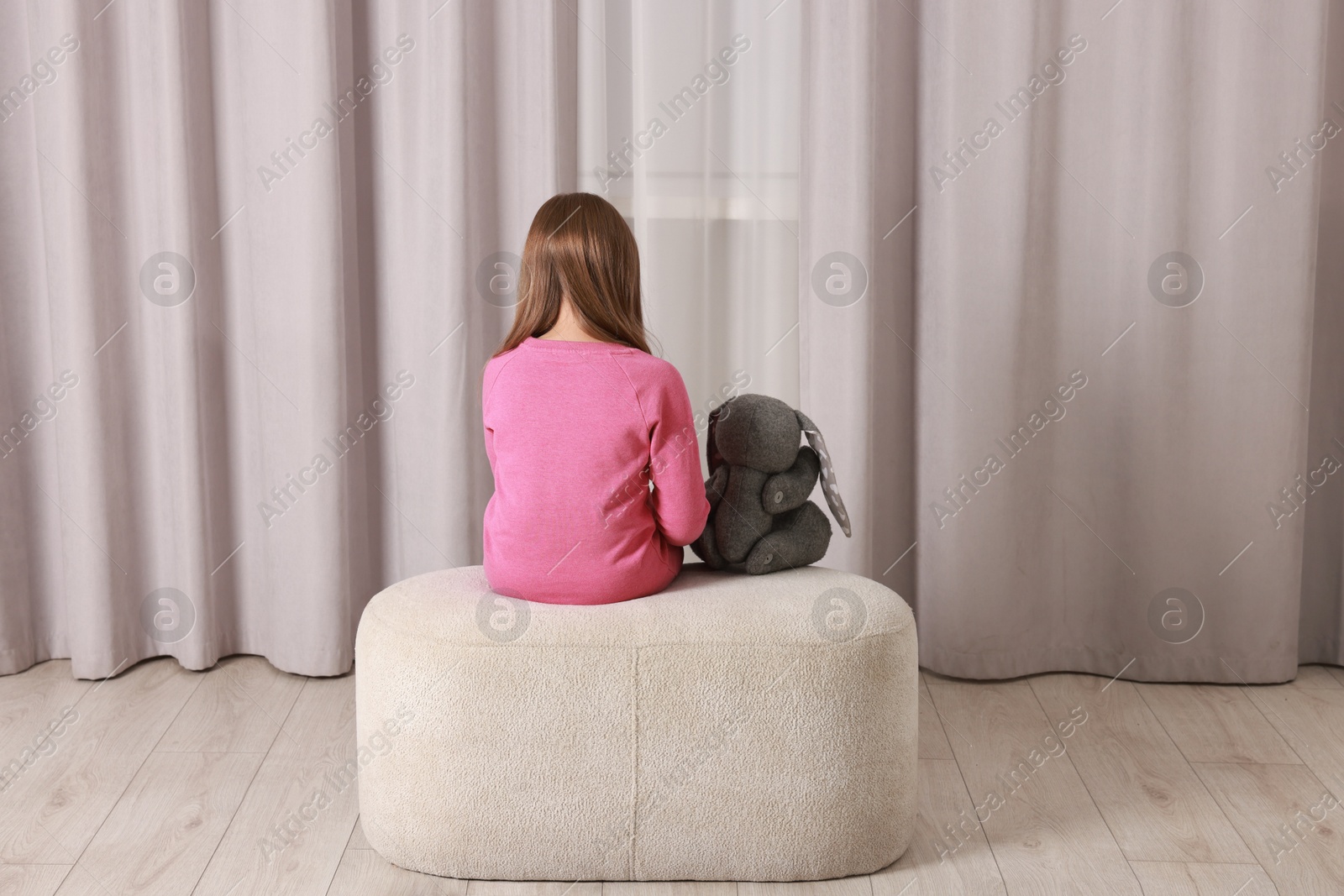 Photo of Autism concept. Lonely little girl with toy bunny on pouf at home, back view