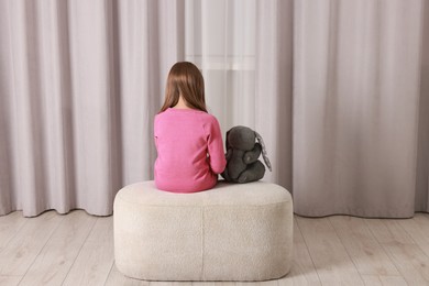 Photo of Autism concept. Lonely little girl with toy bunny on pouf at home, back view