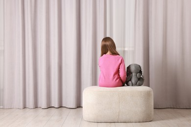 Photo of Autism concept. Lonely little girl with toy bunny on pouf at home, back view. Space for text