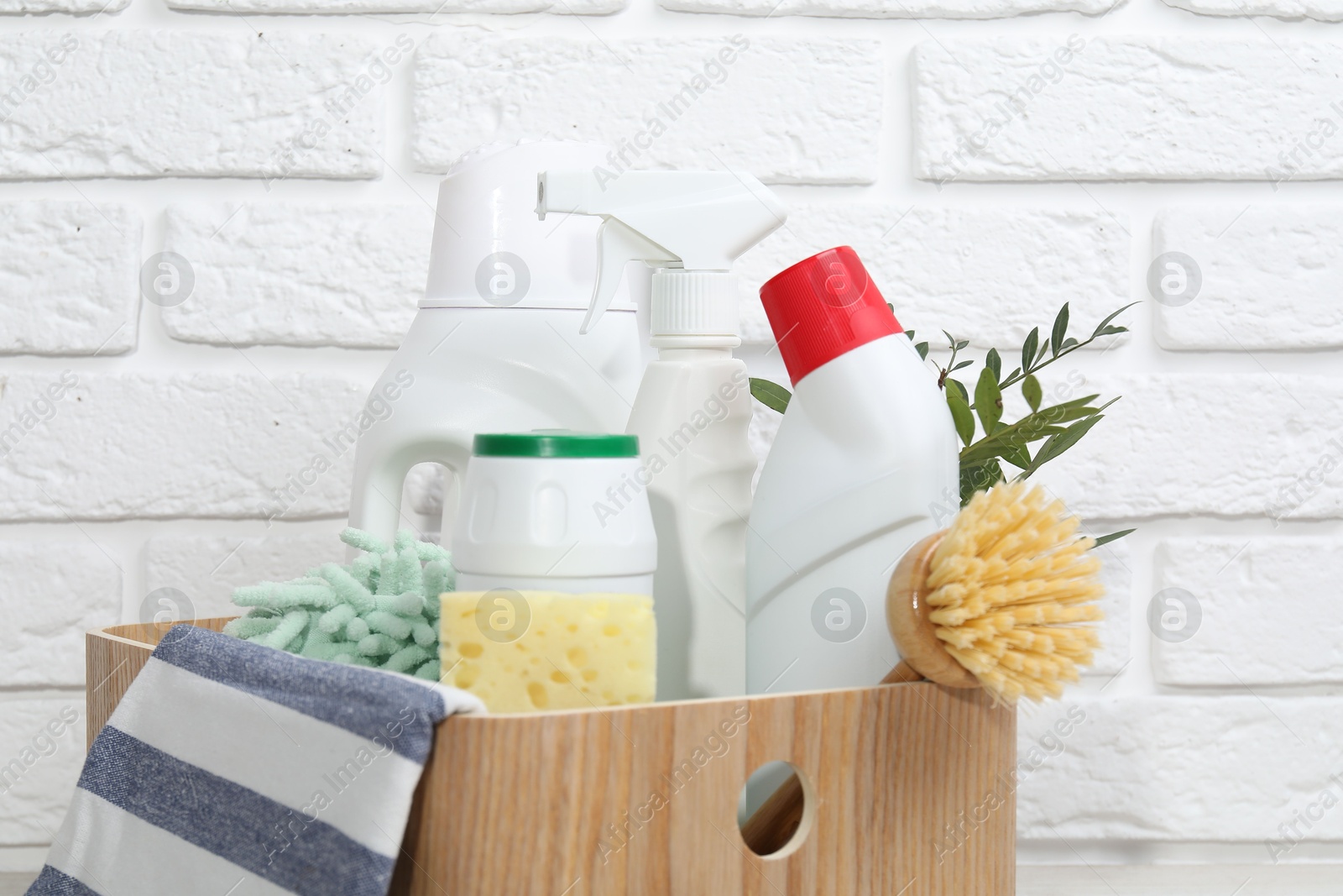 Photo of Eco-friendly cleaning products, supplies and green branches in wooden crate against white brick wall, closeup