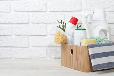 Photo of Eco-friendly cleaning products, supplies and green branches in crate on light wooden table against white brick wall, space for text