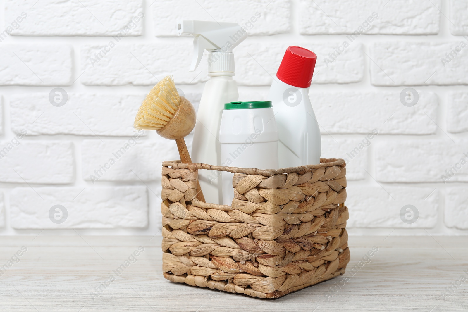 Photo of Eco-friendly cleaning products and brush in wicker basket on wooden table against white brick wall