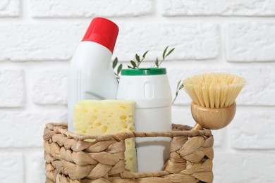 Photo of Eco-friendly cleaning products, supplies and green branches in wicker basket against white brick wall, closeup