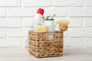 Photo of Eco-friendly cleaning products, supplies and green branches in wicker basket on wooden table against white brick background