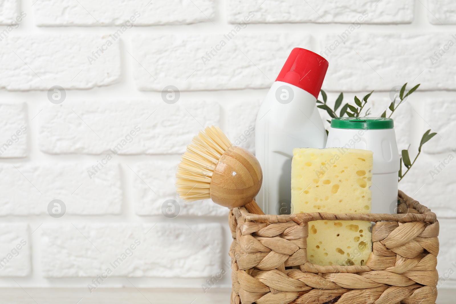 Photo of Eco-friendly cleaning products, supplies and green branches in wicker basket against white brick background, space for text