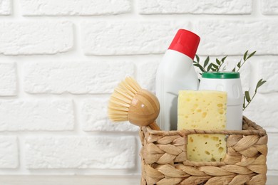 Photo of Eco-friendly cleaning products, supplies and green branches in wicker basket against white brick background, space for text