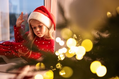 Photo of Little girl with Santa hat near window in room decorated for Christmas