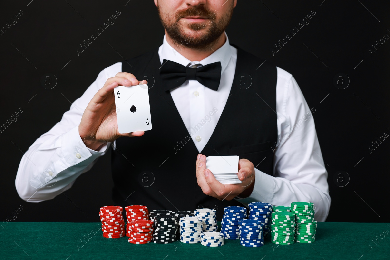 Photo of Professional croupier with playing cards at gambling table against black background, closeup