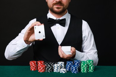 Professional croupier with playing cards at gambling table against black background, closeup