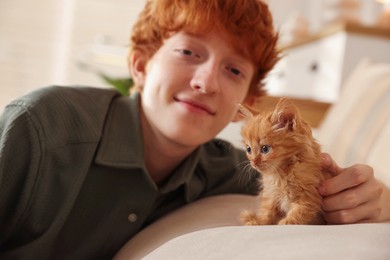 Photo of Redhead teenage boy with cute ginger kitten on sofa indoors, selective focus