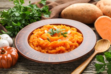 Photo of Tasty mashed sweet potato with pumpkin seeds in bowl, spoon, fresh vegetables and parsley on wooden table, closeup