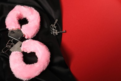 Photo of Pink fluffy handcuffs, keys and black fabric on red background, space for text