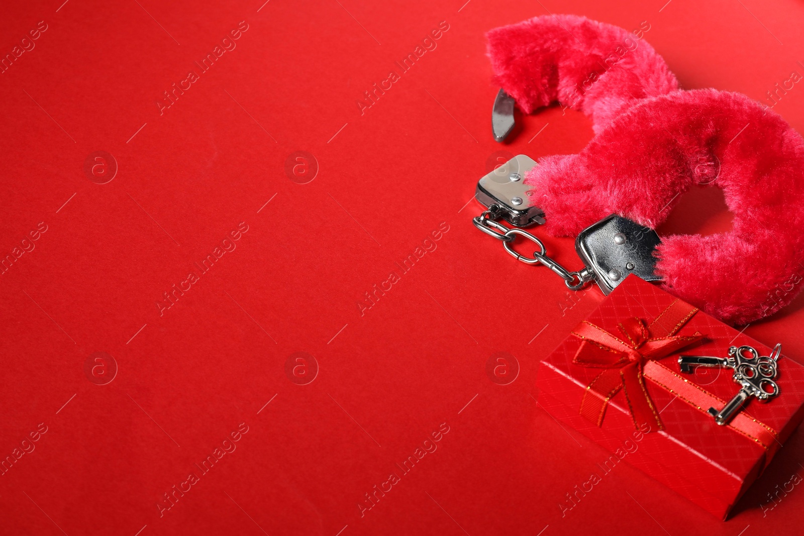 Photo of Bright fluffy handcuffs, keys and gift box on red background, space for text