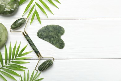 Photo of Face roller, gua sha tool, massage stones and palm leaves on white wooden background, flat lay. Space for text