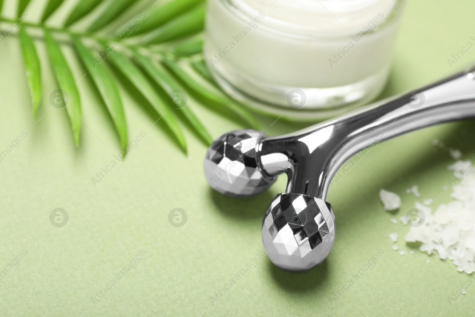 Photo of Metal face roller, cosmetic product, sea salt and palm leaf on green background, closeup