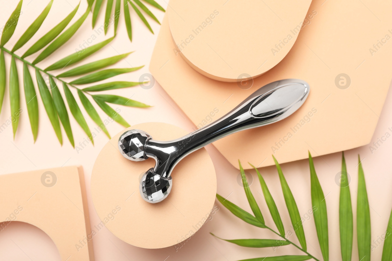 Photo of Metal face roller and palm leaves on beige background, flat lay