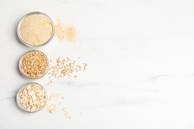 Photo of Oat bran, grains and flakes in bowls on white marble table, flat lay. Space for text