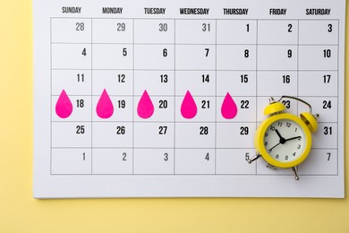 Photo of Menstruation. Calendar with marked dates and alarm clock on beige background, top view