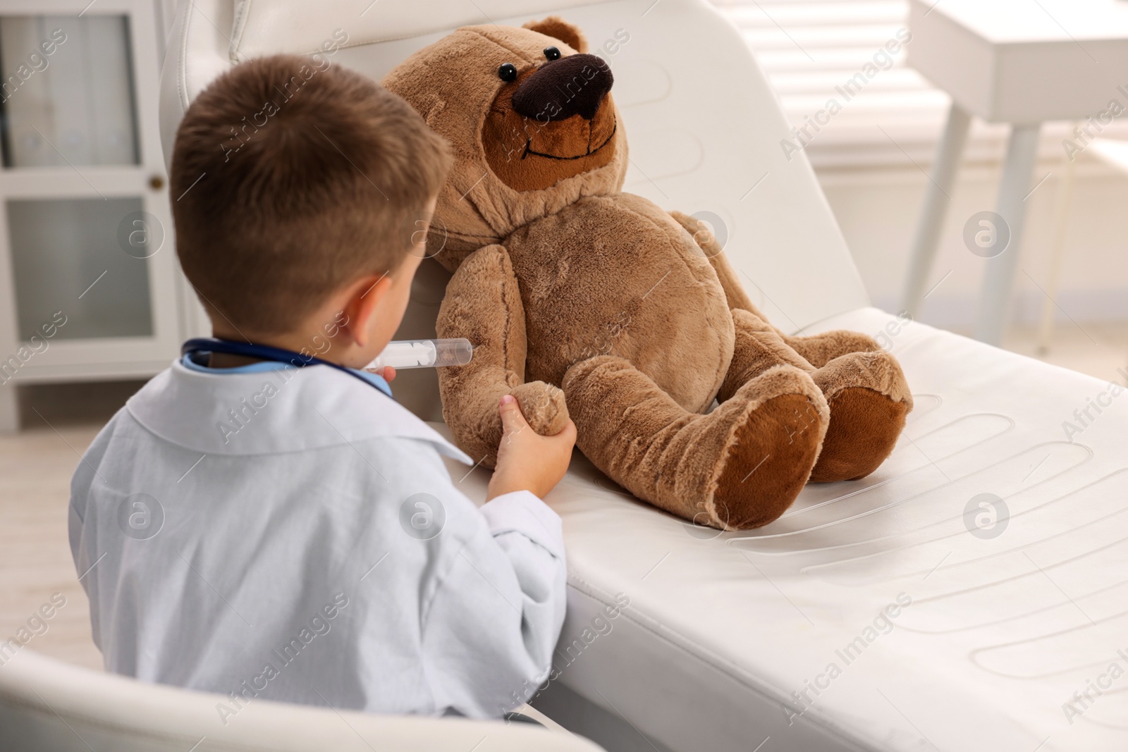 Photo of Little boy with stethoscope, syringe and toy pretending to be doctor indoors, back view