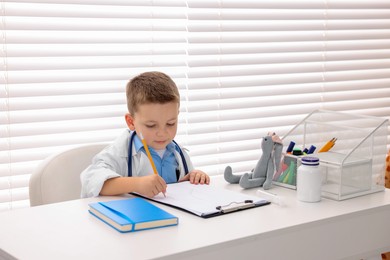 Photo of Little boy with stethoscope and clipboard pretending to be doctor at white table indoors