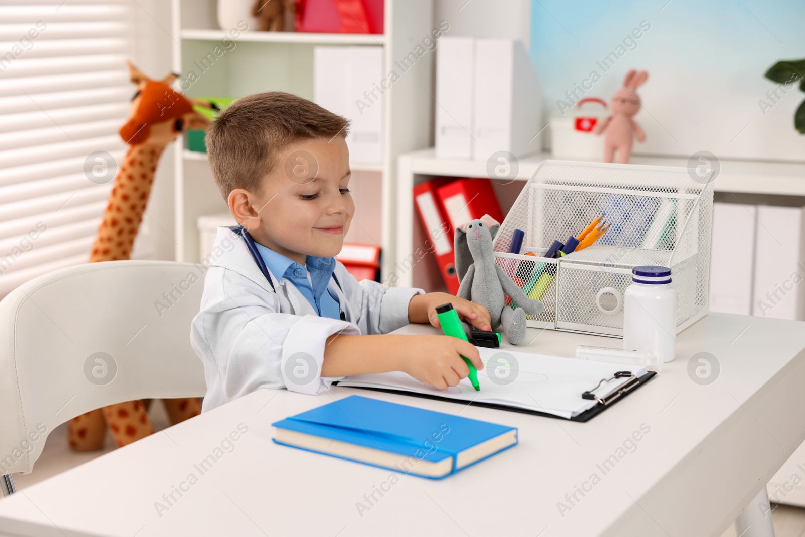 Photo of Little boy with stethoscope and clipboard pretending to be doctor at white table indoors