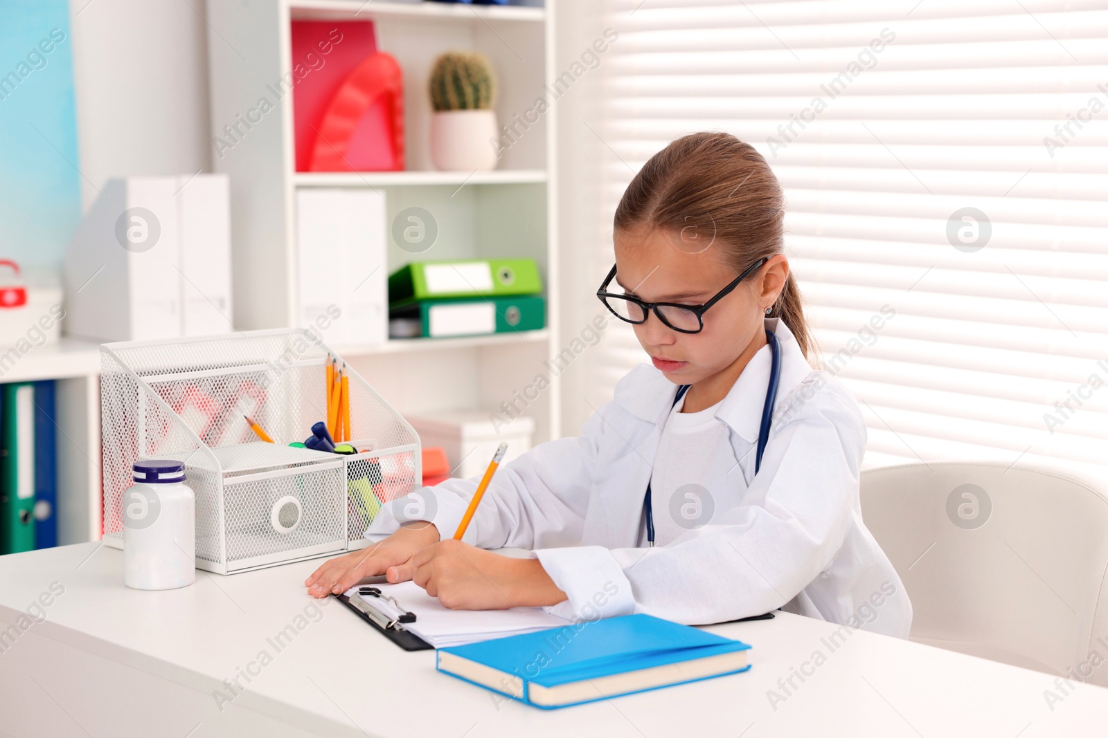 Photo of Little girl with stethoscope and clipboard pretending to be doctor at white table indoors