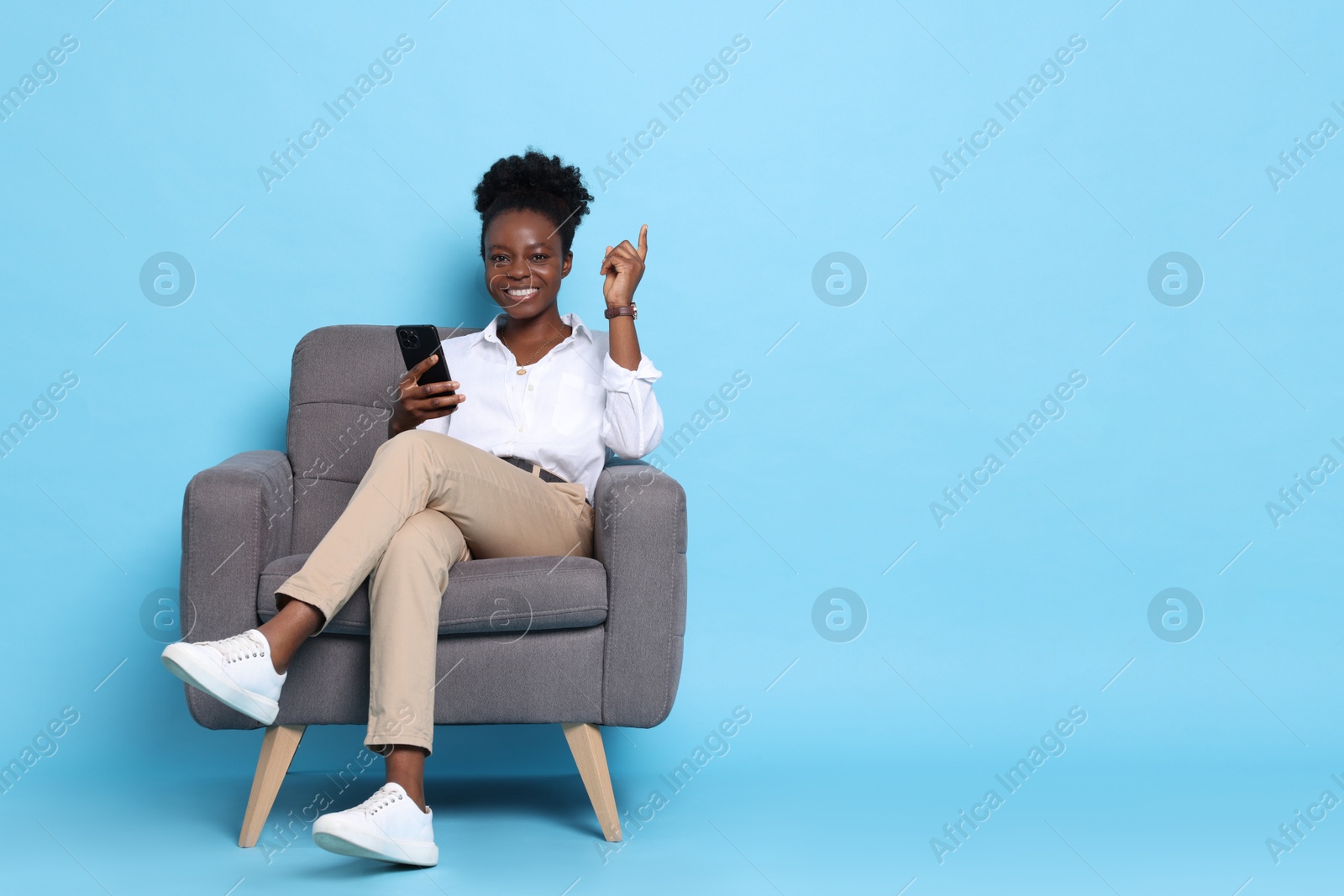 Photo of Cheerful woman with smartphone sitting in armchair and pointing at something on light blue background. Space for text