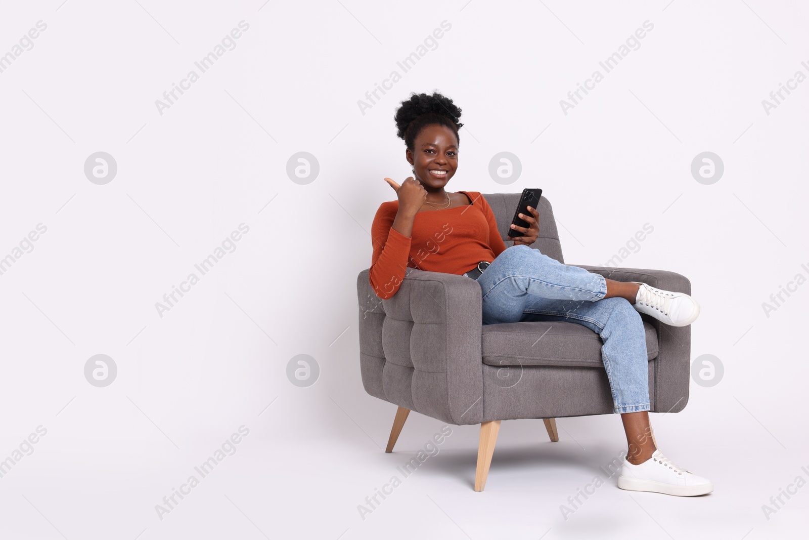 Photo of Cheerful woman with smartphone sitting in armchair and pointing at something on white background. Space for text