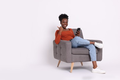 Cheerful woman with smartphone sitting in armchair and pointing at something on white background. Space for text