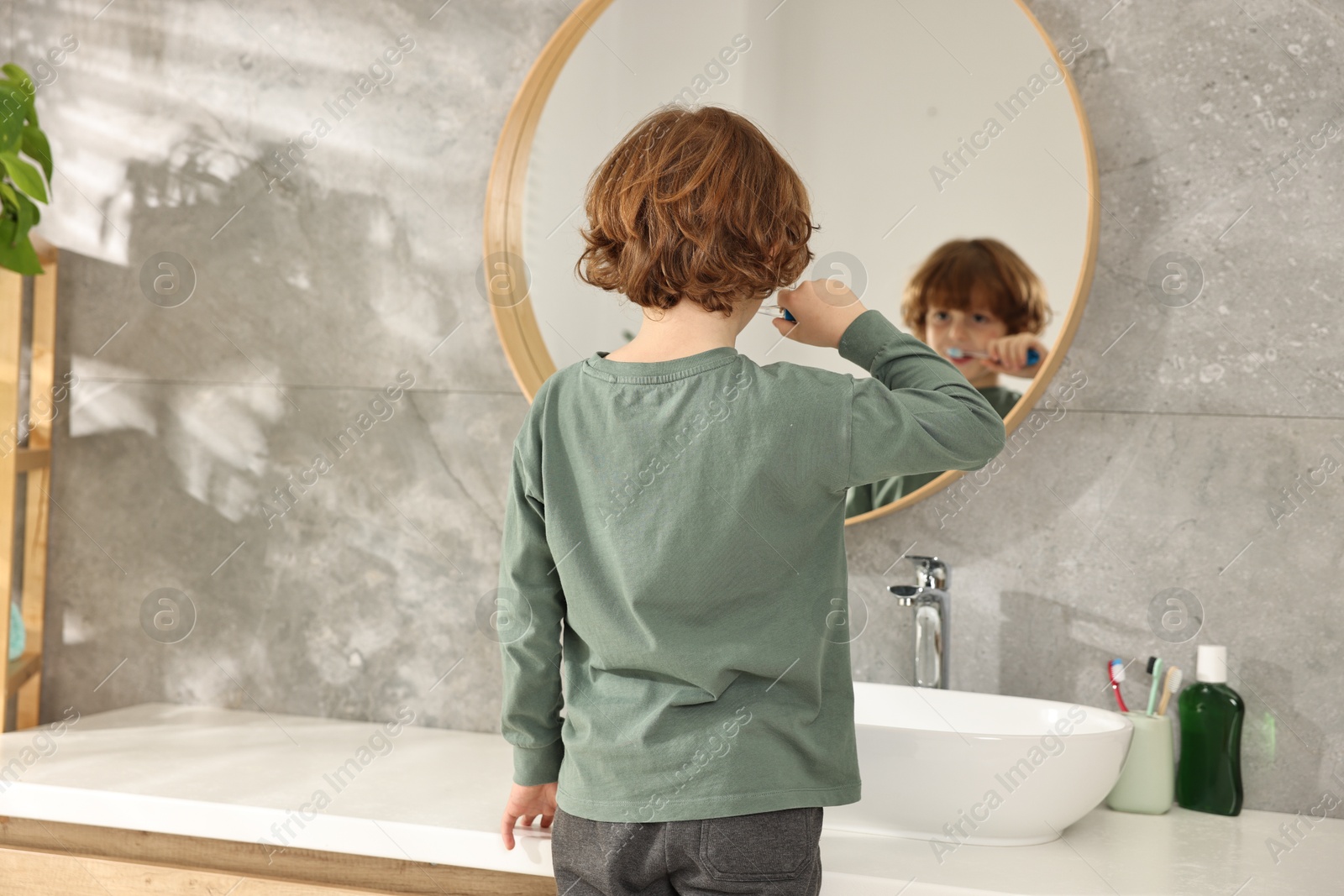 Photo of Cute boy brushing his teeth near mirror in bathroom, back view